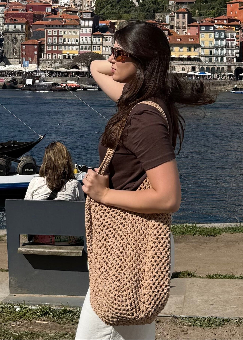 Woman with a handmade crocheted woven tote bag standing by a waterfront with a cityscape in the background