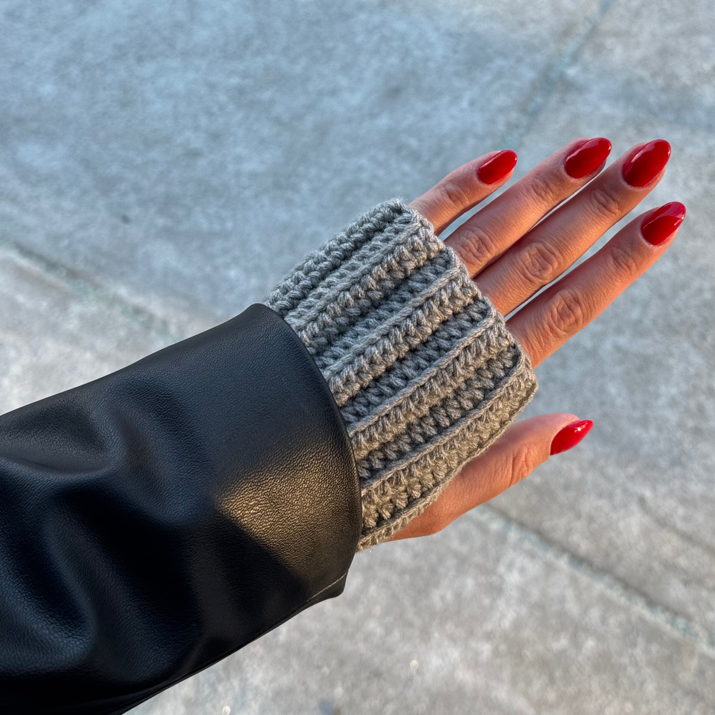 Hand wearing a gray knitted fingerless glove with red nail polish on a concrete surface.